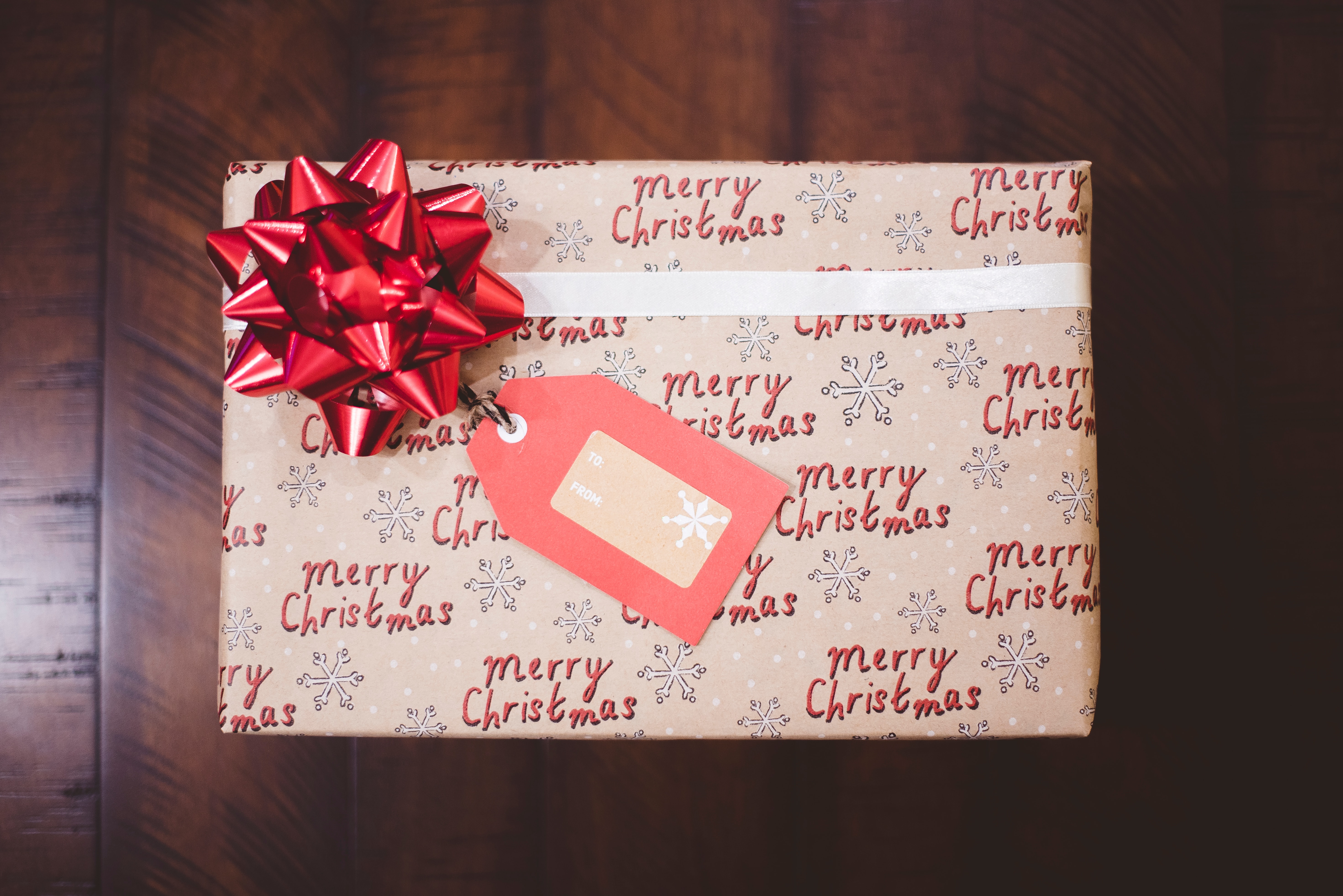 Image shows a rectangluar present with Merry Christmas printed across the wrapping paper. It has a red bow on the top left hand corner.