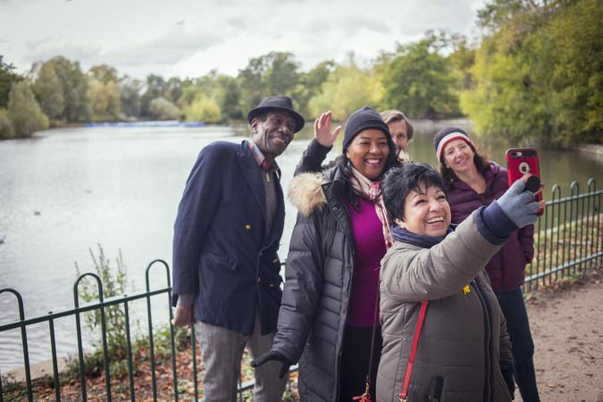 A group of older adults, dressed warmly and standing beside a lake, smiling into a camera for a selfie