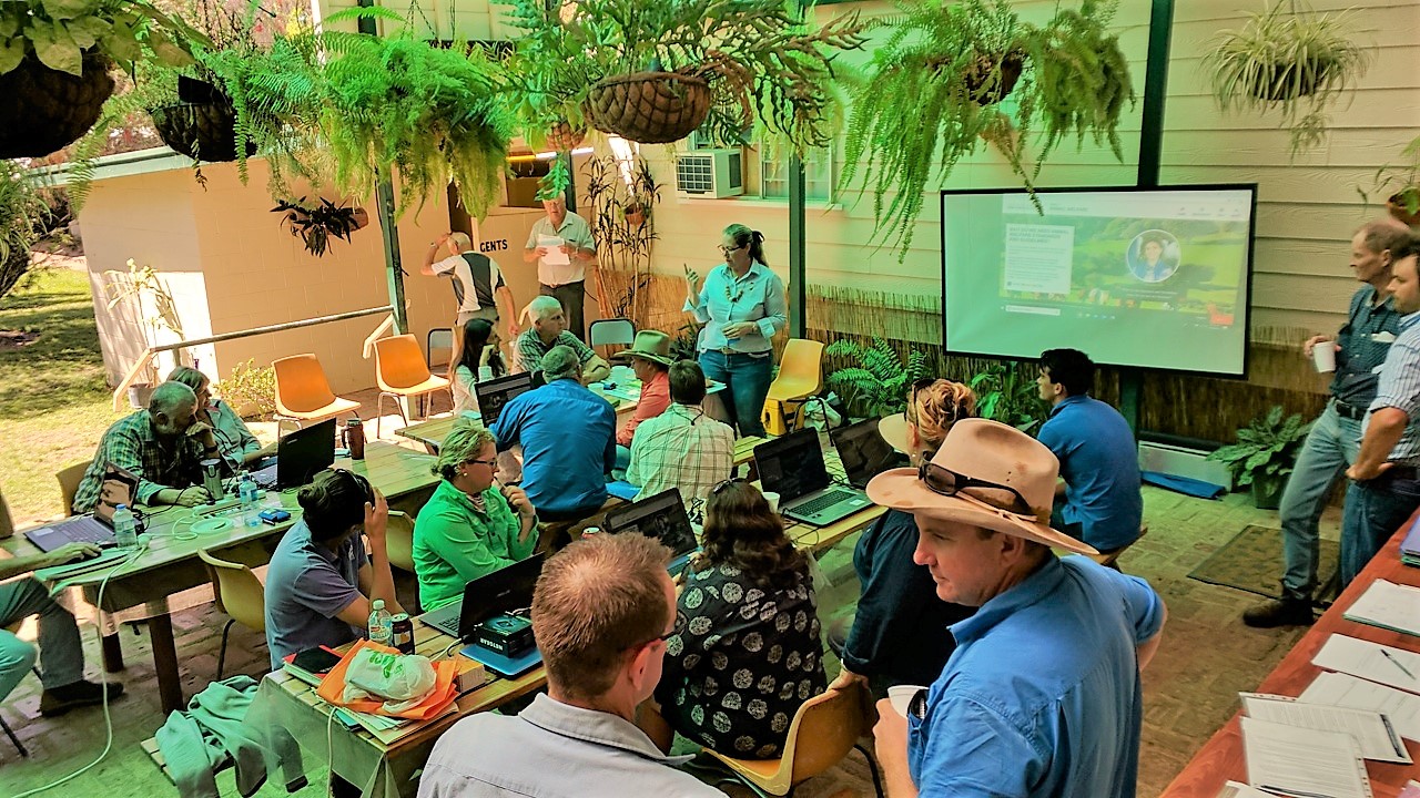 overhead view of farmers sitting & standing, chatting. Screen in front of them. 