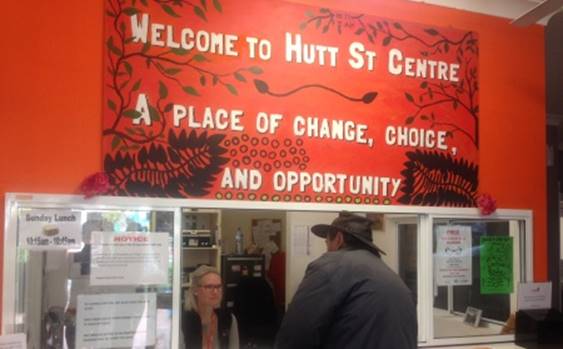 Image of back of client speaking with staff member behind counter, under a red banner reading "Welcome to Hutt St Centre. A place of change, choice and opportunity"